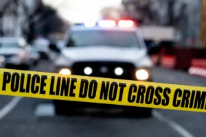A police car with flashing lights is seen behind a "POLICE LINE DO NOT CROSS CRIME" tape at a crime scene.