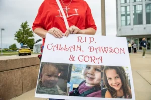 The photo shows a person holding a sign with the text "R.I.P. CALLAN, DAWSON & CORA" and images of three children, indicating a memorial or tribute.