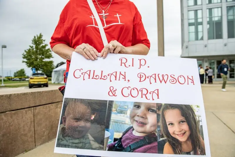 The photo shows a person holding a sign with the text "R.I.P. CALLAN, DAWSON & CORA" and images of three children, indicating a memorial or tribute.