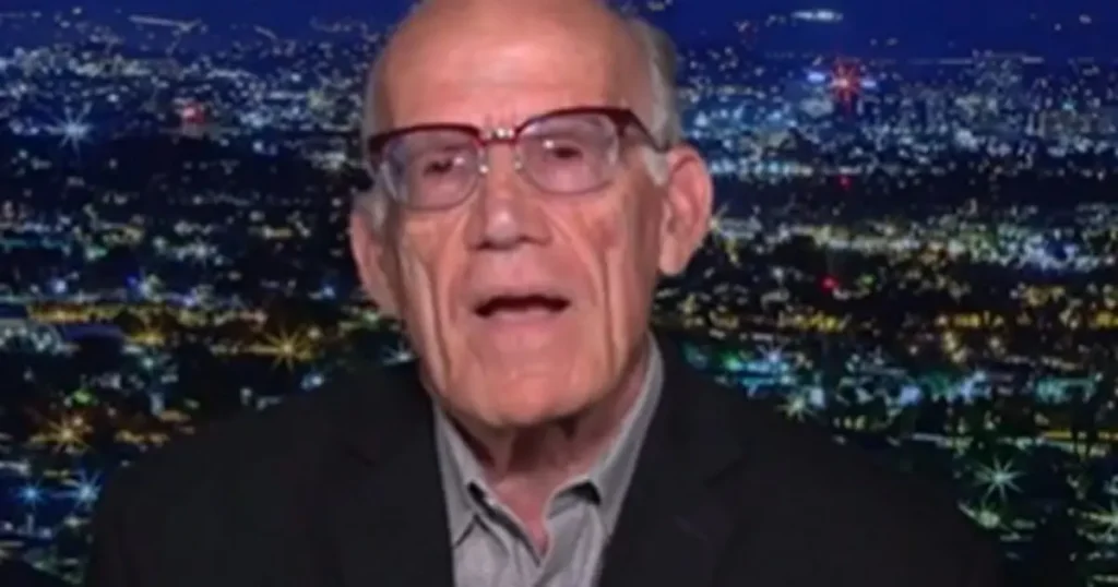 The image shows an elderly man wearing glasses and a suit, set against a backdrop of a cityscape at night.