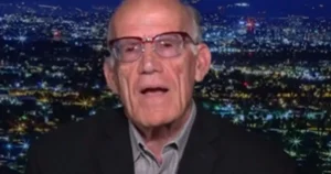 The image shows an elderly man wearing glasses and a suit, set against a backdrop of a cityscape at night.