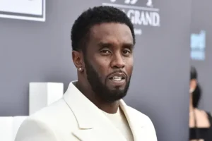 The image shows a man in a white suit at what appears to be a formal event, possibly a red carpet or awards ceremony.