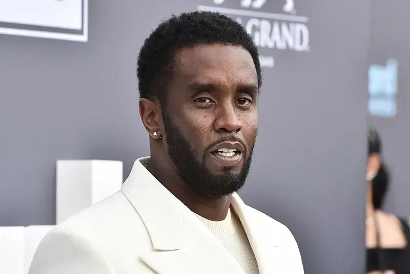 The image shows a man in a white suit at what appears to be a formal event, possibly a red carpet or awards ceremony.