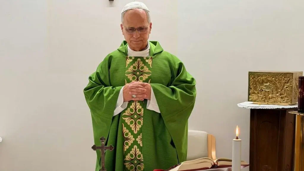 A priest in a green vestment is seen in a prayerful pose, with a Bible and a candle on the altar in front of him.