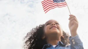 A young child holds an American flag, smiling and looking up, symbolizing patriotism and national pride.