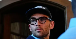 The image shows a person wearing a black cap with the word "DEMOCRATIC" on it and glasses, standing in front of a wooden door.