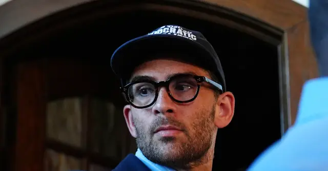 The image shows a person wearing a black cap with the word "DEMOCRATIC" on it and glasses, standing in front of a wooden door.