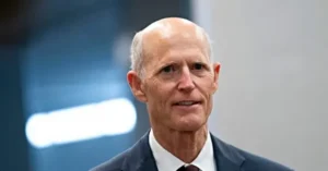 The image shows a man in a suit and tie, appearing to be in a professional or formal setting.