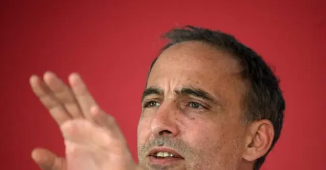 The image shows a man gesturing with his hand while speaking, set against a red background.