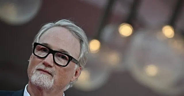 The image shows a man with gray hair and a beard, wearing glasses and a suit, set against a blurred background with bokeh lights.