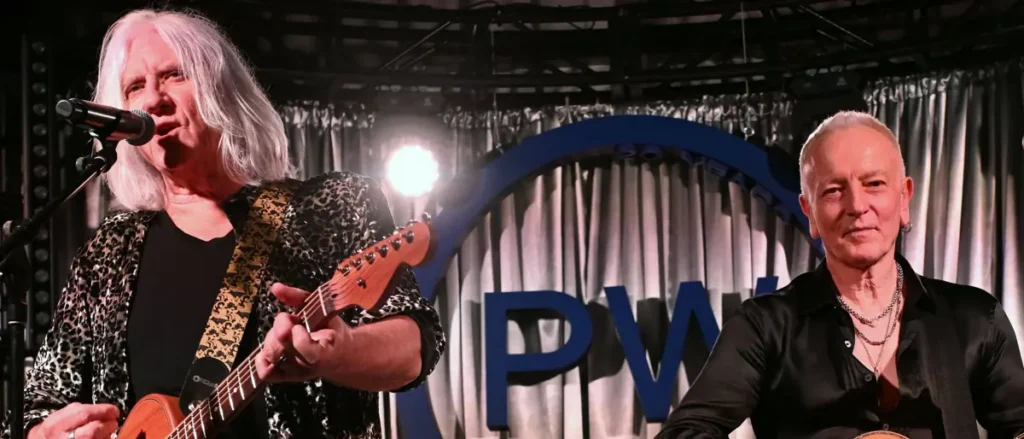 Two musicians perform on stage, one playing guitar and the other seated, with a backdrop featuring the letters "PWI."