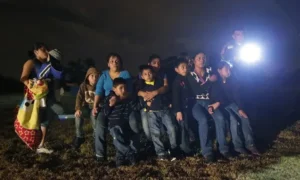 A group of people, including children, are gathered outdoors at night, illuminated by a flashlight, suggesting a moment of shared experience or concern.