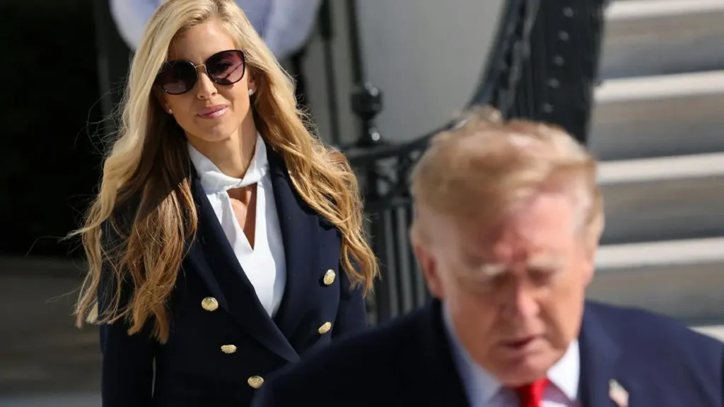 The image shows a woman in a navy blue blazer and sunglasses walking past a man in a suit.