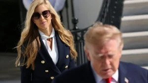 The image shows a woman in a navy blue blazer and sunglasses walking past a man in a suit.