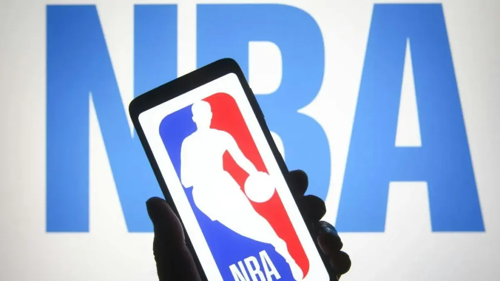 A hand holds a smartphone displaying the NBA logo in front of a large NBA sign.