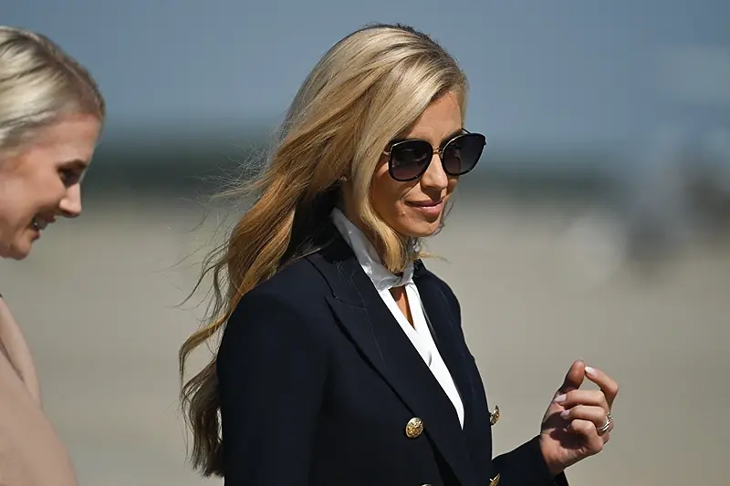 The photo shows a woman in a black blazer and sunglasses walking outdoors, with another person partially visible on the left side.
