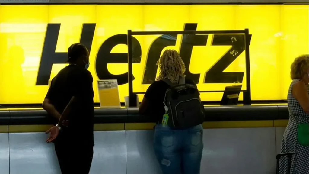 A group of people stands in front of a bright yellow Hertz sign, possibly at an airport or rental car location.