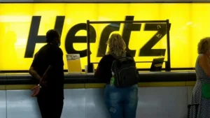 A group of people stands in front of a bright yellow Hertz sign, possibly at an airport or rental car location.