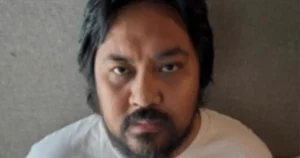 The image shows a person with dark hair and a beard, wearing a light-colored shirt, against a plain background.