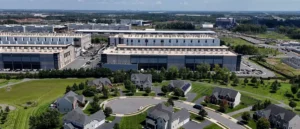 The image shows an aerial view of a suburban neighborhood with residential houses surrounded by greenery, adjacent to a large industrial complex with multiple buildings and parking lots.
