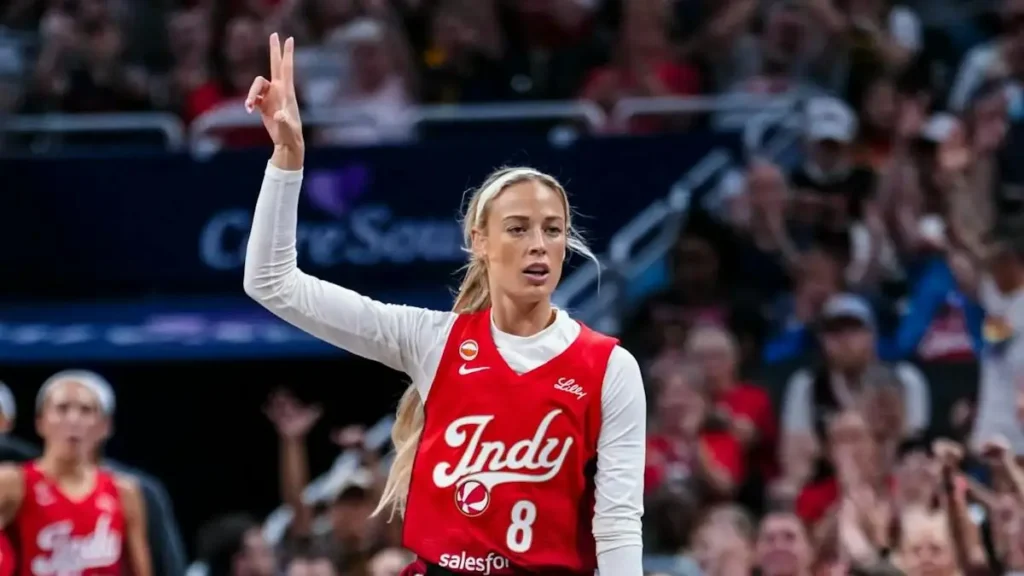 A basketball player in a red jersey with "Indy" and the number 8 raises her hand in a gesture of celebration or signaling during a game.