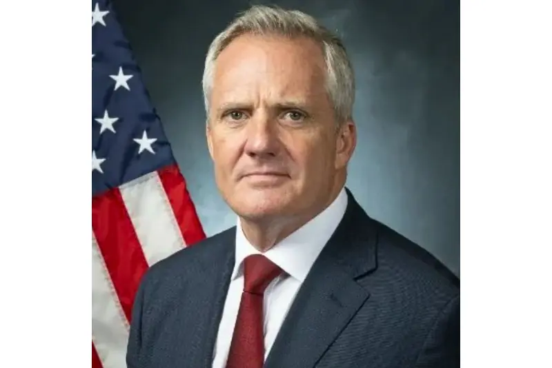 The image shows a man in a suit and tie standing in front of an American flag.