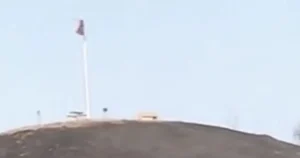 A flag flies atop a hill, with a structure visible in the foreground.
