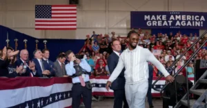 A man in a white outfit walks down a stage at a rally with a crowd and an "MAKE AMERICA GREAT AGAIN!" sign in the background.