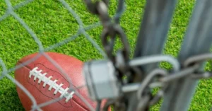 A football lies on a grassy field behind a chain-link fence.