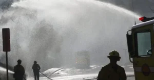 Firefighters are seen managing a large water spray from a fire truck, likely as part of a firefighting operation.