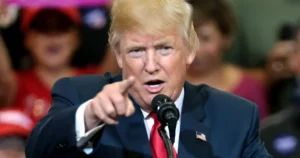 The image shows a person in a suit and tie, speaking into a microphone and pointing forward, with a crowd in the background.