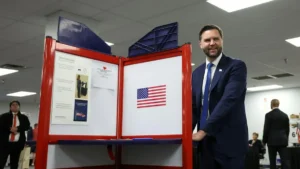 A man in a suit stands next to a voting booth with an American flag, smiling at the camera.