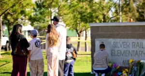 A group of people gather at a memorial at Silver Rail Elementary School, where flowers and a small pumpkin are placed.