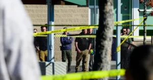 Police officers are seen behind a fence and yellow crime scene tape at an outdoor location.