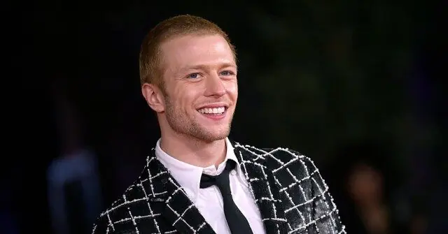 The image shows a person smiling and wearing a stylish black and white checkered blazer over a white shirt.