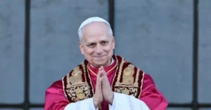 The image shows a person in papal attire, with hands clasped in prayer, standing in front of a stone wall.