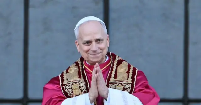 The image shows a person in papal attire, with hands clasped in prayer, standing in front of a stone wall.