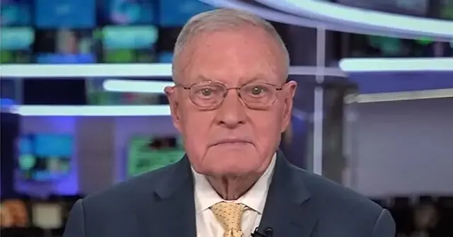 The image shows a man in a suit and tie, likely a news anchor or commentator, in a studio setting with a blurred background of screens and lights.