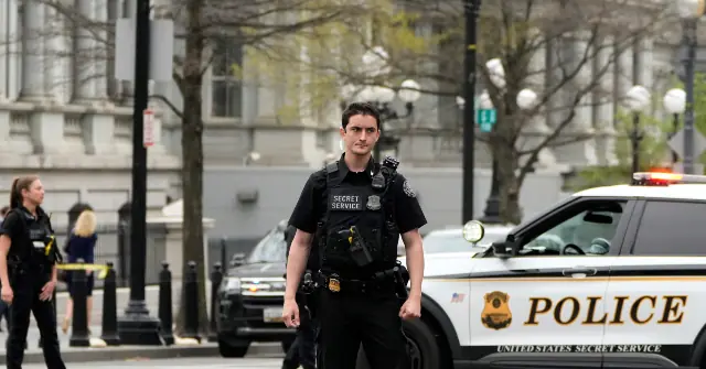 A Secret Service officer stands in front of a police car, with other personnel and vehicles in the background, indicating a security presence in an urban setting.
