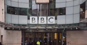 The image shows the exterior of a BBC building with the BBC logo prominently displayed on the facade.