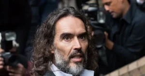 The image shows a man with long hair and a beard, surrounded by photographers and cameras, suggesting a public or media event.