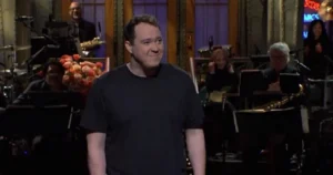 The image shows a man in a black shirt standing in front of a band and a floral arrangement, likely during a performance or event.