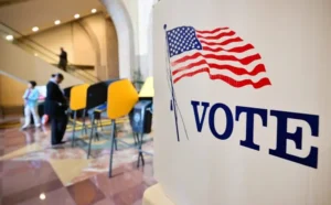 The image shows a voting booth with an American flag and the word "VOTE" prominently displayed, set in a public space with people in the background.