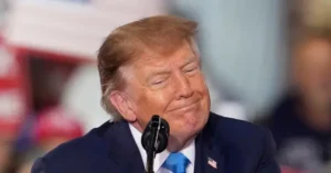 The image shows a man in a suit and tie speaking into a microphone at what appears to be a public event or rally.