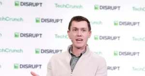 The image shows a person speaking at a TechCrunch Disrupt event, as indicated by the repeated logos in the background.