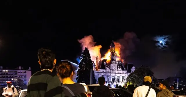 A crowd of people watches as a building is engulfed in flames and smoke at night.
