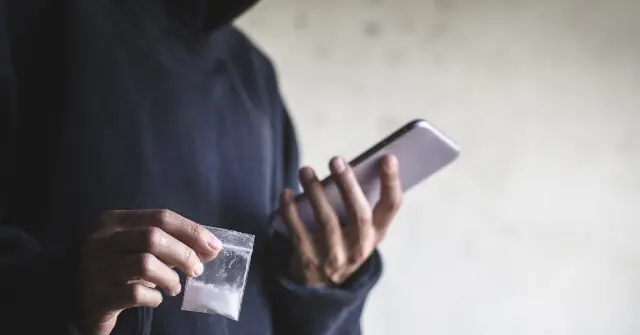The image depicts a person holding a smartphone in one hand and a small plastic bag containing a white substance in the other.