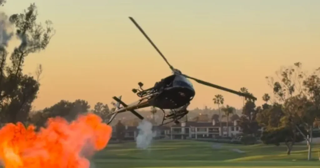 A helicopter is seen flying low over a golf course with a bright orange flare in the foreground, set against a backdrop of trees and buildings under a warm sky.