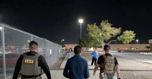 The image shows law enforcement officers, including those with "HSI" and "FB" vests, walking along a fenced area at night, with a person in a blue shirt and another in casual attire nearby.
