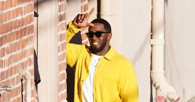 A person wearing a bright yellow jacket and sunglasses is making a peace sign with their hand while standing against a brick wall.